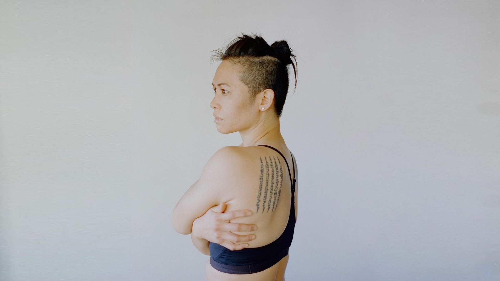 Profile portrait of Katty Strite with arms crossed, Sak Yant shoulder tattoo visible against a pale wall
