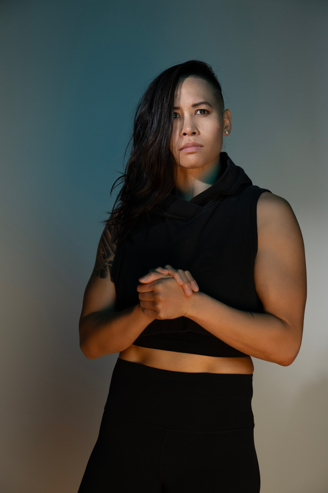 Katty Strite in a sleeveless black hoodie, hands clasped, lit by warm and cool studio light