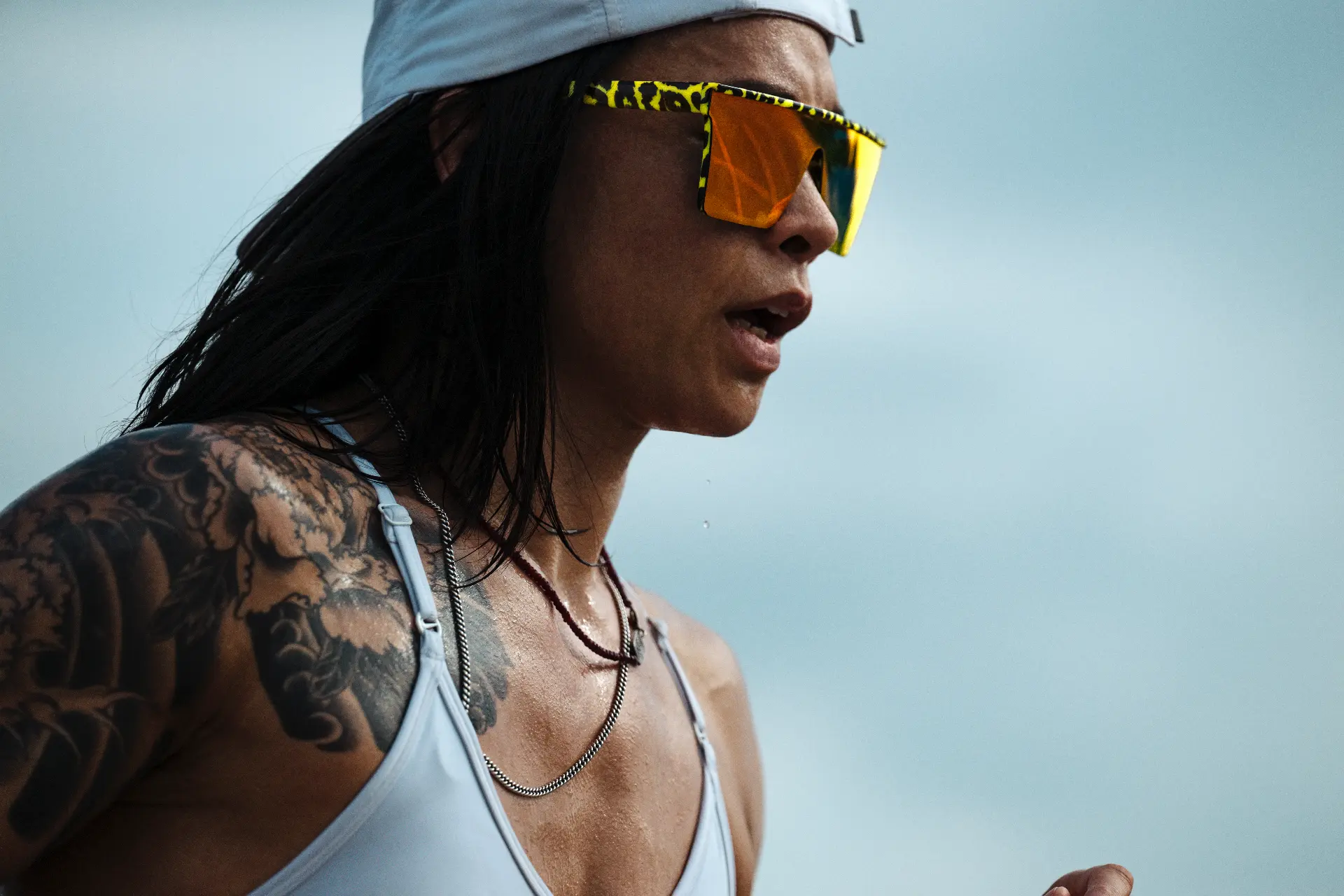 Katty Strite in profile wearing a light cap and reflective sunglasses, tattoos on her shoulder